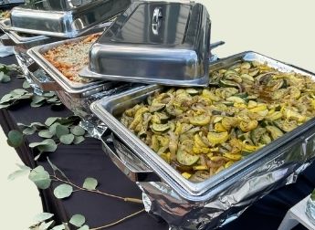 A catering line of stainless chafing dishes on a black-draped table with eucalyptus garland, the front tray filled with grilled zucchini and yellow squash