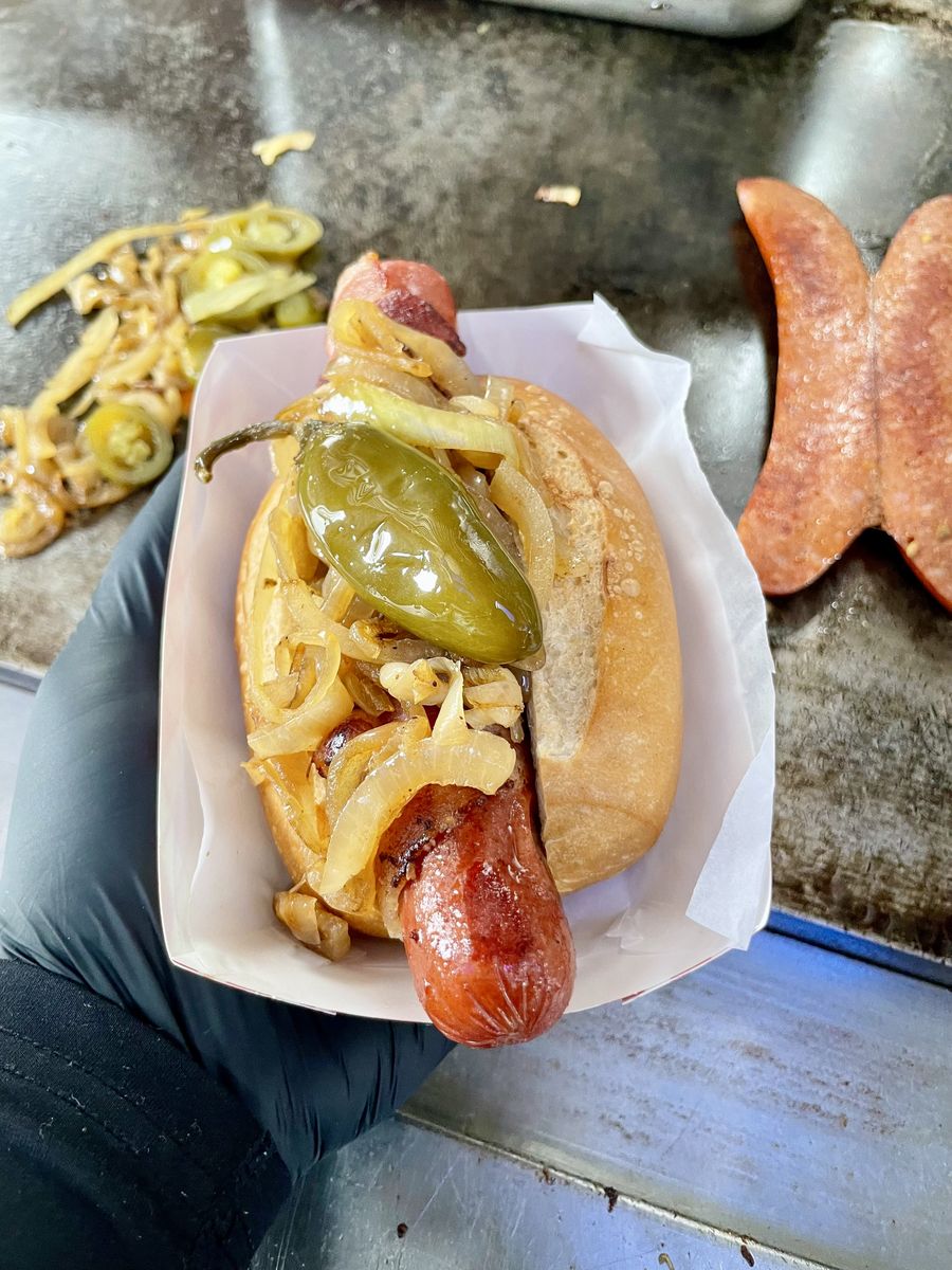 A gloved hand holding a red sausage on a roll with grilled onions and a whole pickled pepper
