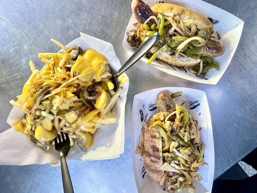 Two sausages on rolls loaded with grilled onions and green peppers, next to cheese fries with peppers and onions
